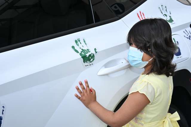 A young girl wearing a protective mask stands near a car with painted handprints on its side.