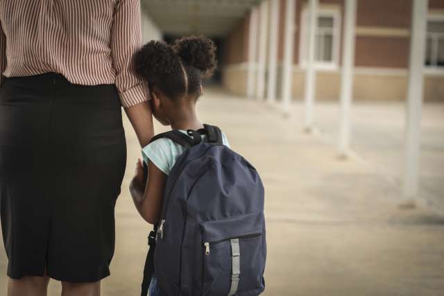 A young girl wearing a backpack leans on her mother's arm as they walk toward school.