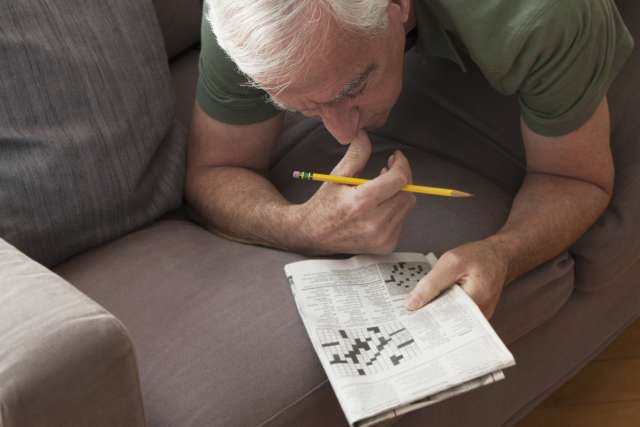 An older man does a crossword puzzle while lying on the couch