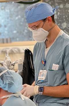 Dr. K. Elliott Higgins III, M.D. stands to the right side of the frame facing the left in scrubs with a surgical cal and mask. 