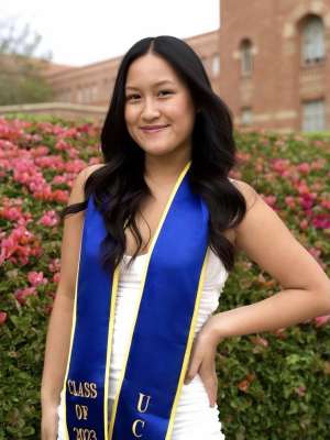 A woman with wavy black hair, wearing a white dress and graduation sash, posing on UCLA campus