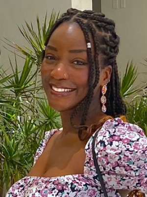 A woman with long braided hair, wearing a floral dress and earrings, smiling in front of a forested background