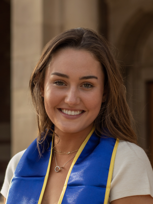 A woman with blonde hair, wearing her UCLA sash, smiling on UCLA campus
