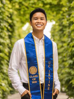 A man with short black hair, wearing a white collared shirt with his UCLA sash, posing in front of a forested background