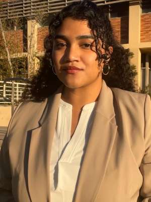A woman with long black curly hair, wearing a beige suit coat with a white shirt, posing in front of UCLA