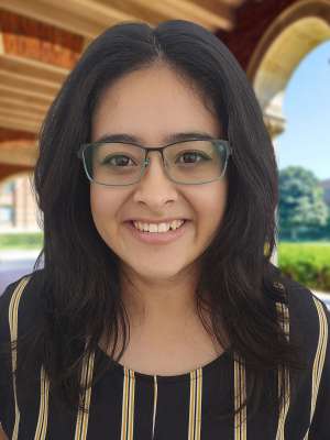 A woman with long black hair, wearing glasses and a striped shirt, smiling on UCLA campus