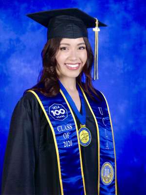 A woman with black curly hair, smiling in her graduation cap, gown, and sash in front of a royal blue background