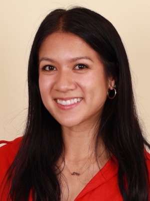 A woman with long black hair, wearing a red shirt and necklace, smiling in front of a blank background
