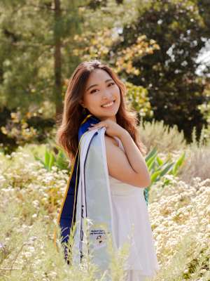 A woman with long brown hair, posing with her graduation sash in a forested setting