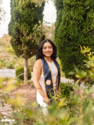 A woman with long black hair, wearing her graduation sash, posing in a forested setting