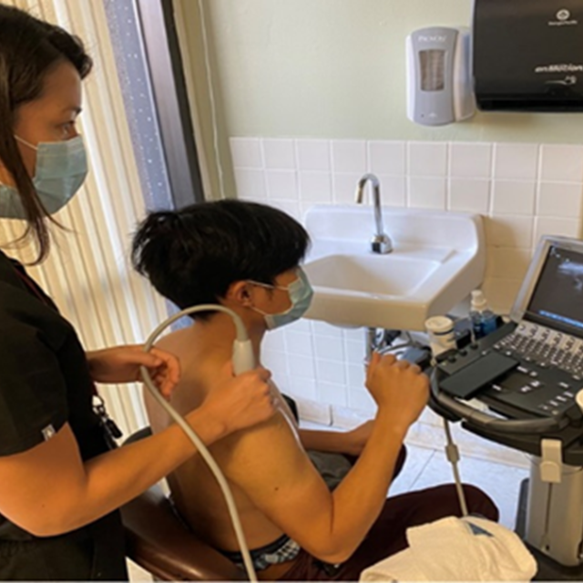 A woman with dark hair is performing an ultrasound procedure on a man's right shoulder